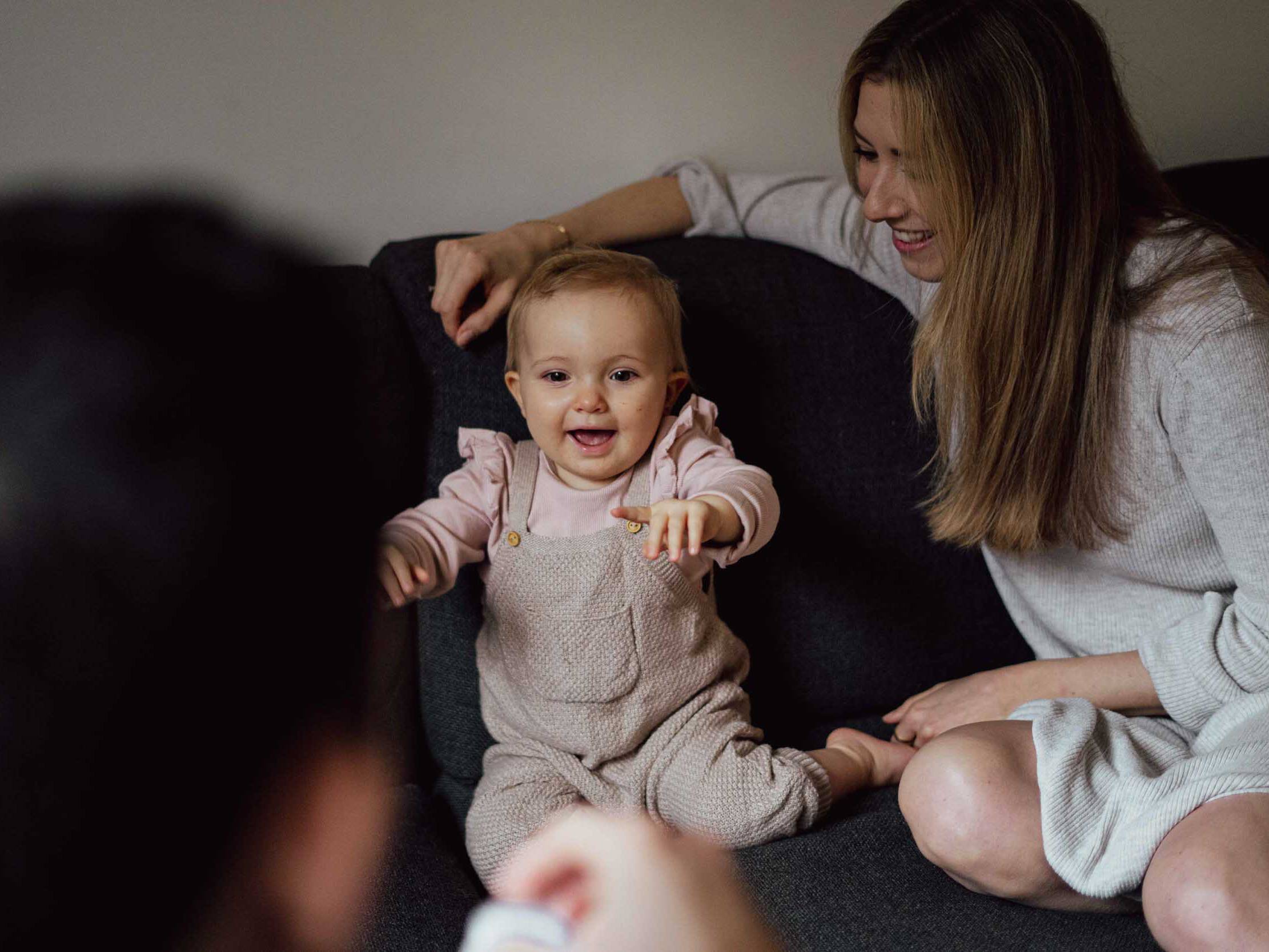 Kleines Kind sitzt mit Mutter auf den Sofa und streckt die Arme zum Papa aus