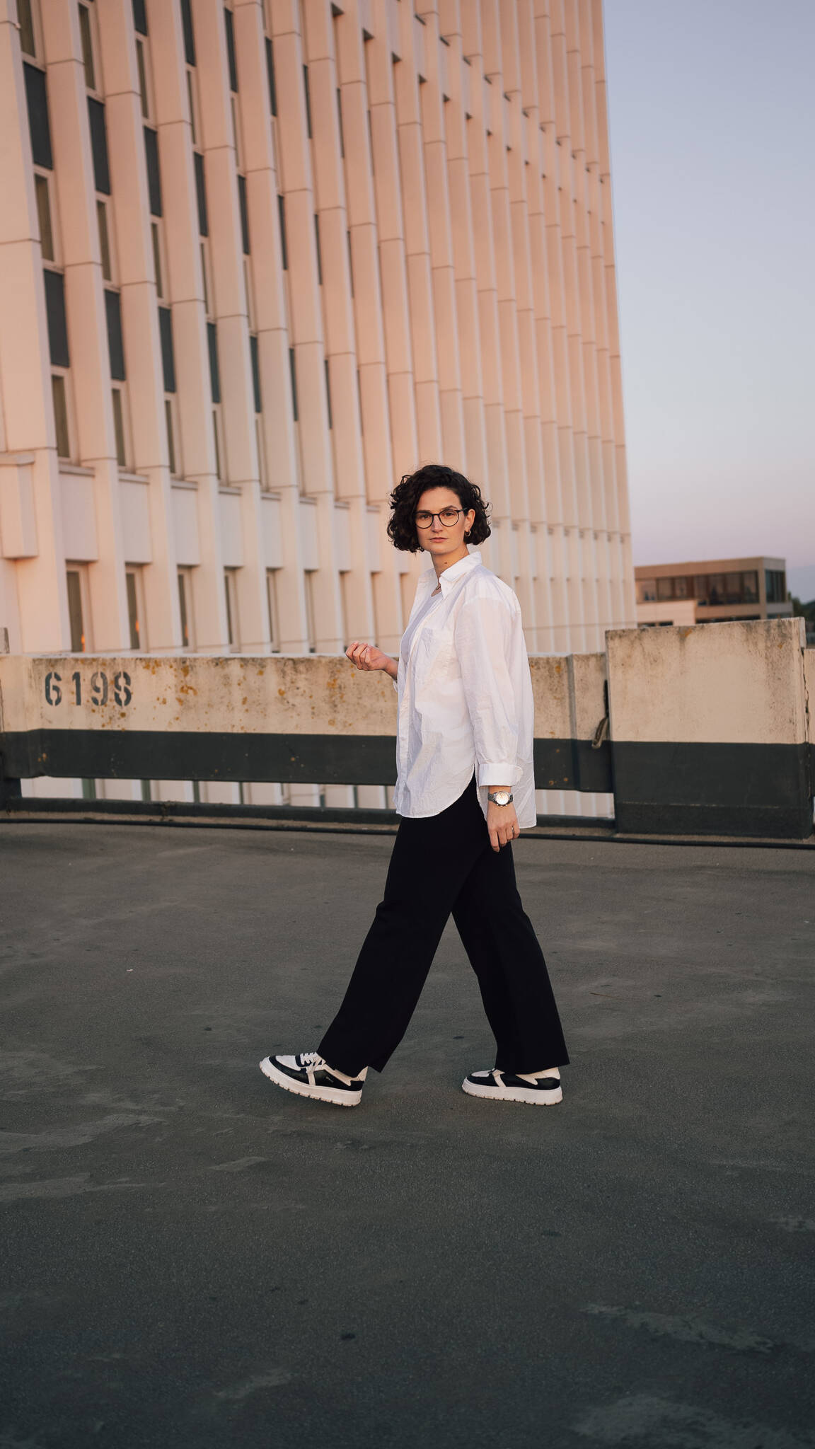 Frau in weißer Bluse und schwarzer Hose auf Parkdeck, Hochhaus im Hintergrund