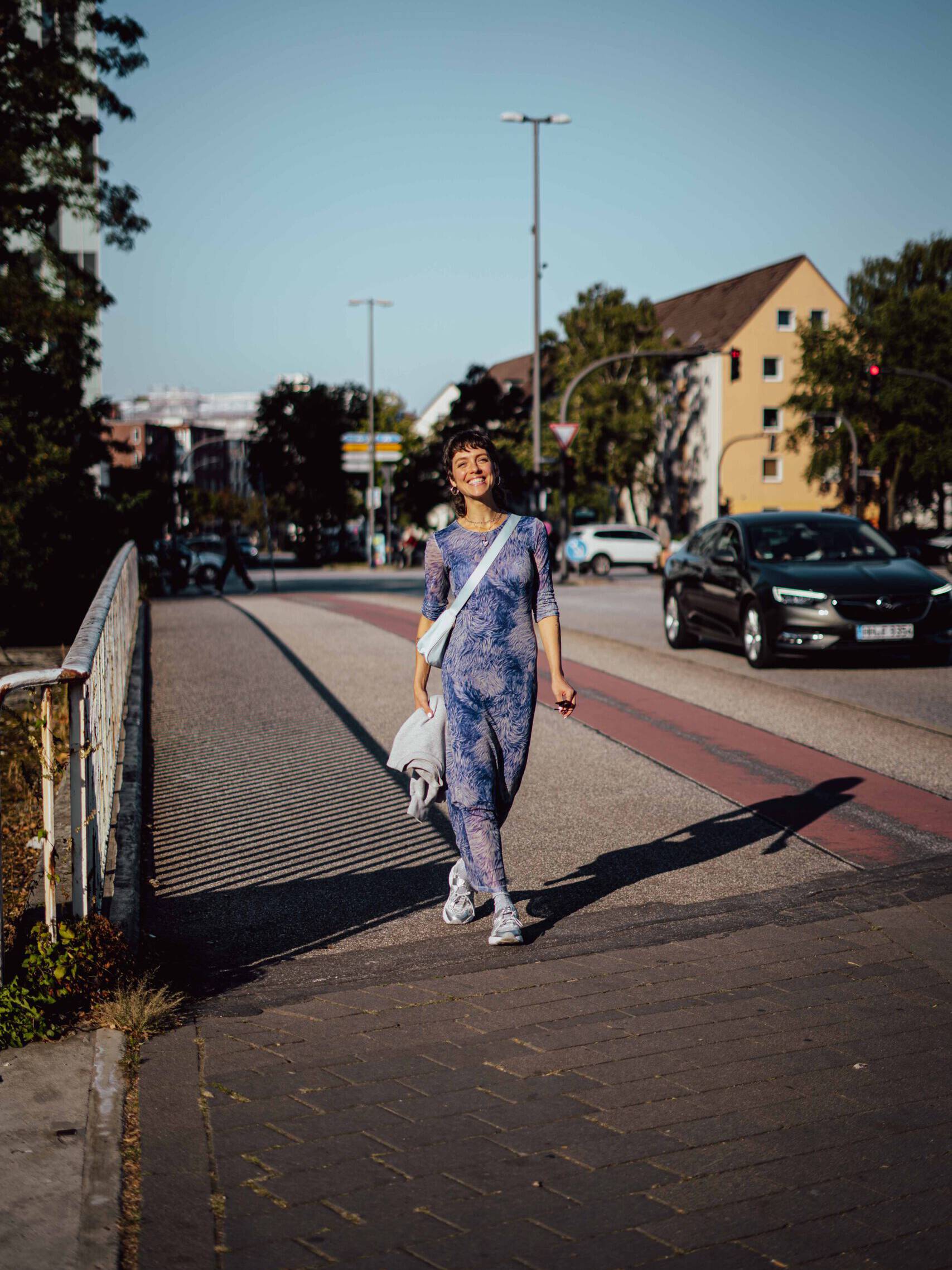 Frau in blaue gemustertem Kleid auf Gehweg, im Hintergrund Ampel und Autos